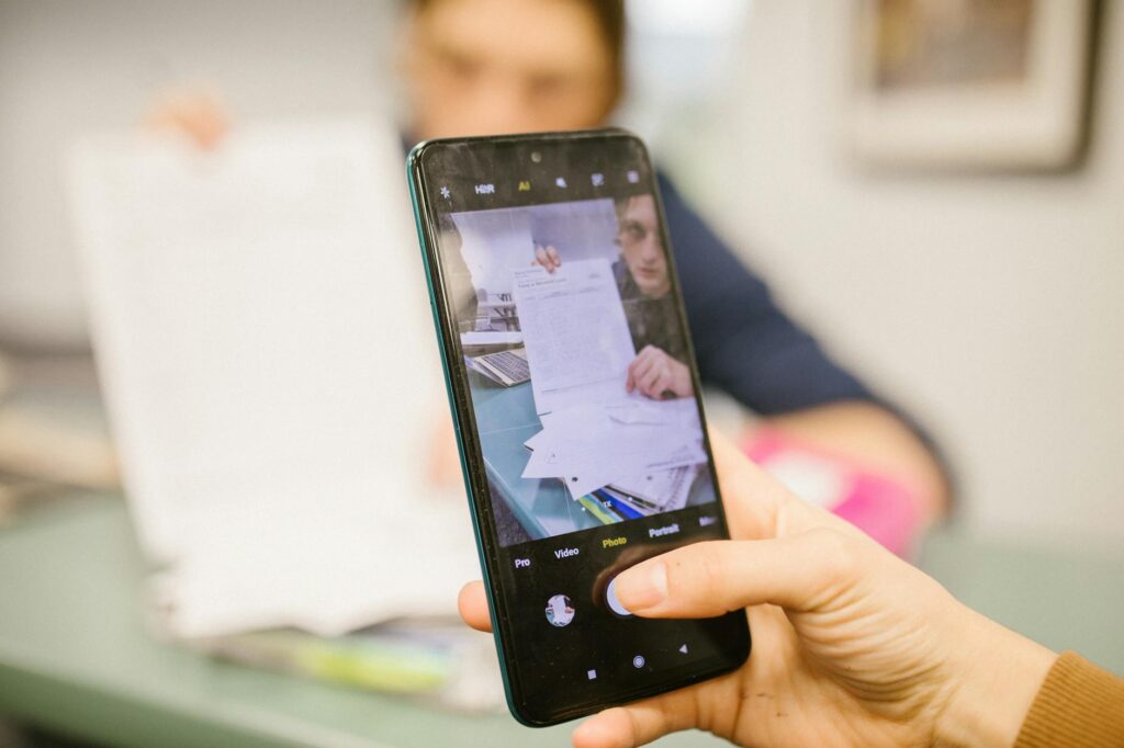 A young student holding a smartphone that displays a two-factor authentication code, with a laptop in the background.