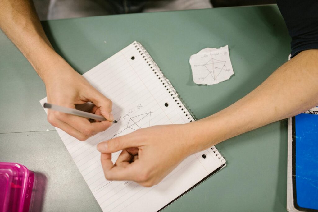 Close-up of a student's hand writing notes in a spiral notebook during a university lecture.