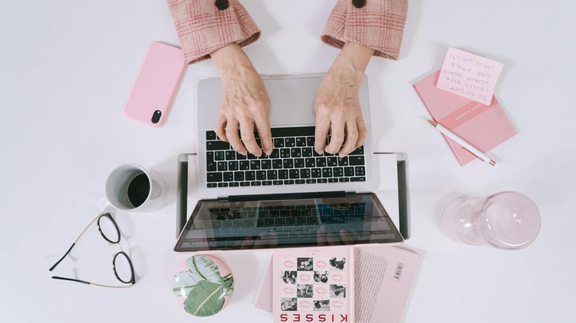 A freelancer smiling while working on a laptop at a sunlit home office desk.