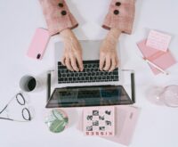 A freelancer smiling while working on a laptop at a sunlit home office desk.