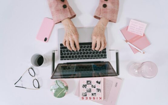 A freelancer smiling while working on a laptop at a sunlit home office desk.