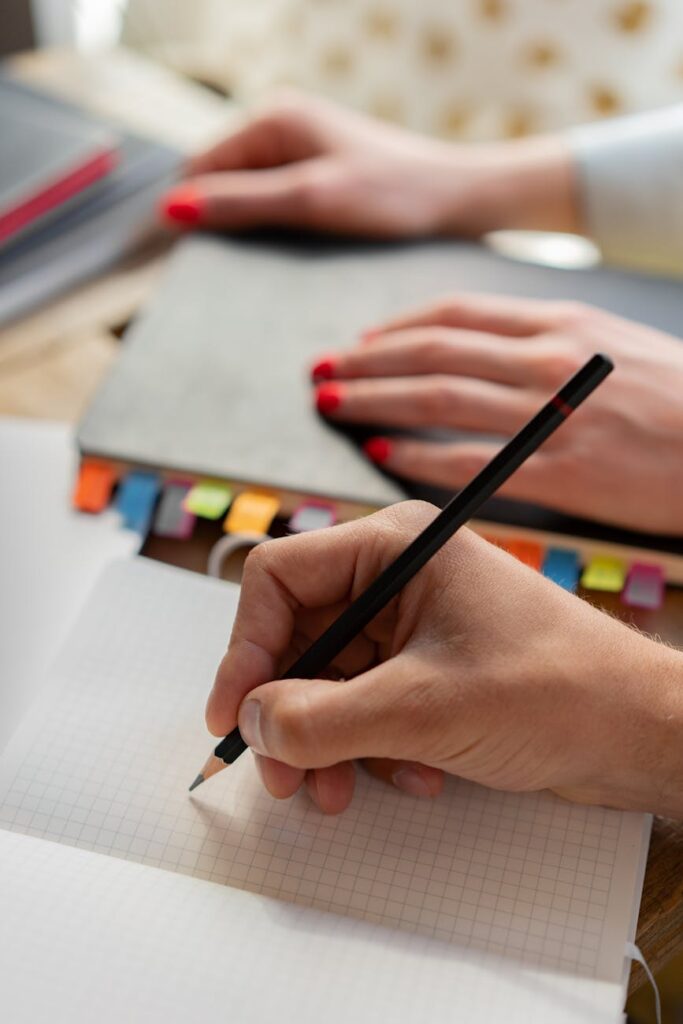 A detailed shot of a student's hand writing notes with a pen in a spiral notebook during a class.