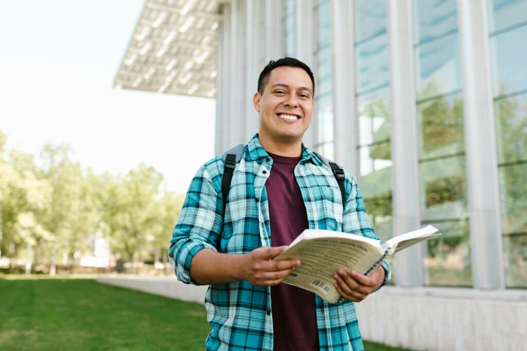 A close-up, friendly portrait of a female university student with a backpack smiling directly at the camera on a green campus.