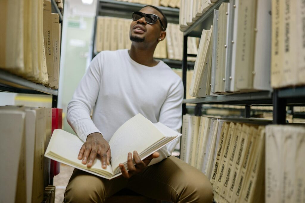 A young student with glasses sits alone in a quiet campus space, looking thoughtfully at their notebook before a difficult conversation.