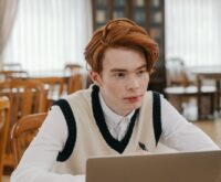 A young student wearing headphones concentrates on their laptop in a sunlit, quiet library.