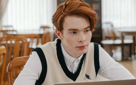 A young student wearing headphones concentrates on their laptop in a sunlit, quiet library.