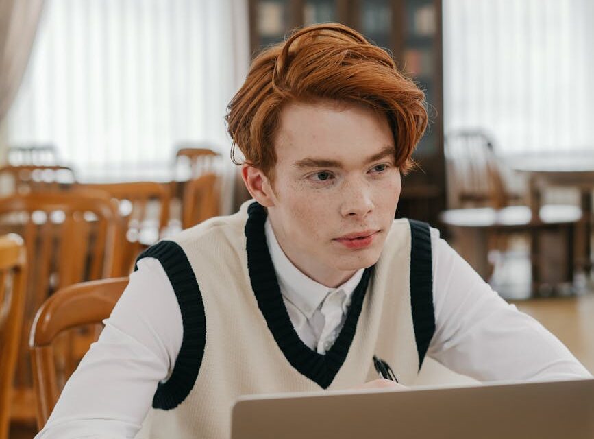 A young student wearing headphones concentrates on their laptop in a sunlit, quiet library.