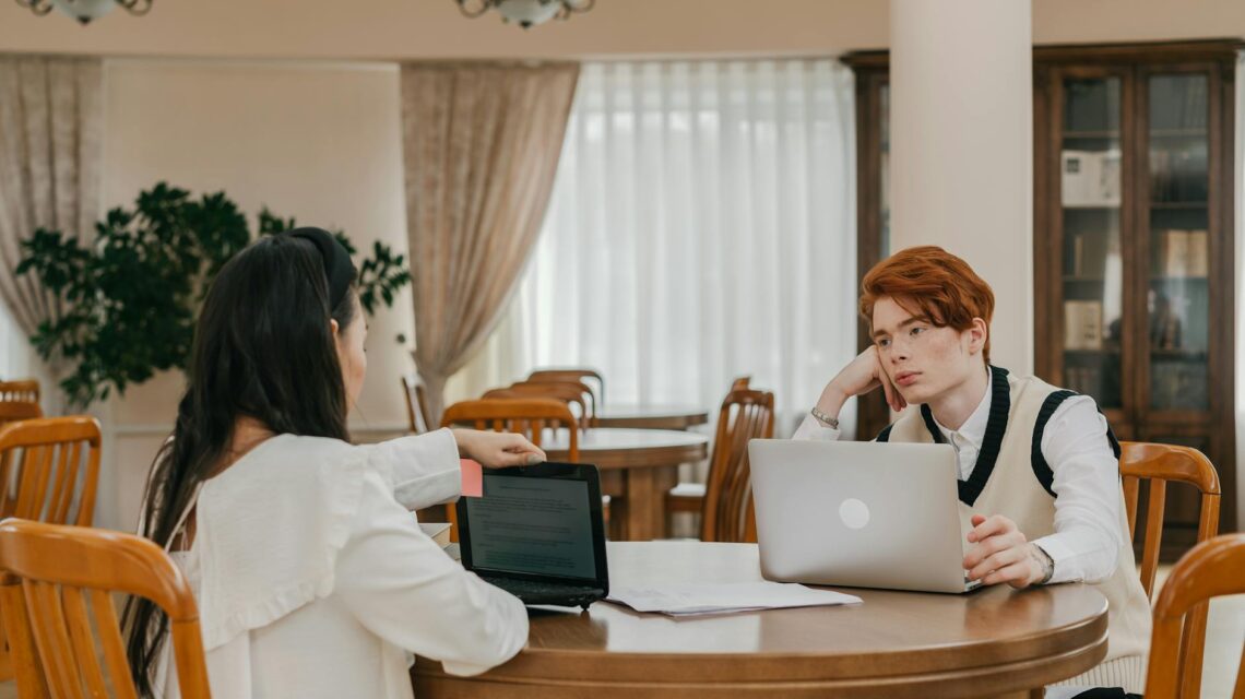 A focused university student with headphones on, studying at a wooden table in a sunlit library.