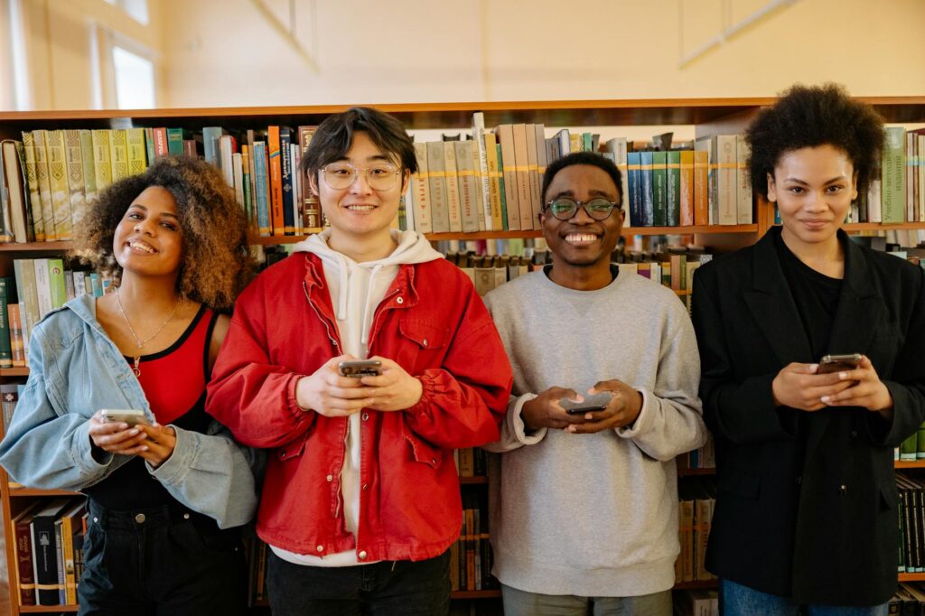 A diverse group of students gathered around a table, smiling and working together on a laptop.