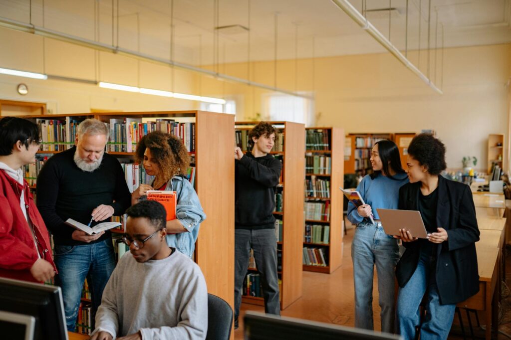 A multicultural group of four students gathered around a laptop, smiling and discussing their work in a modern library.