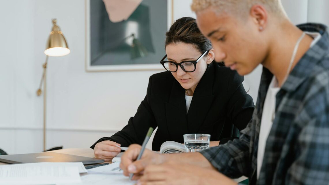 A diverse group of four university students sit around a wooden table in a well-lit library, focused on their books and laptops.