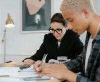 A diverse group of four university students sit around a wooden table in a well-lit library, focused on their books and laptops.