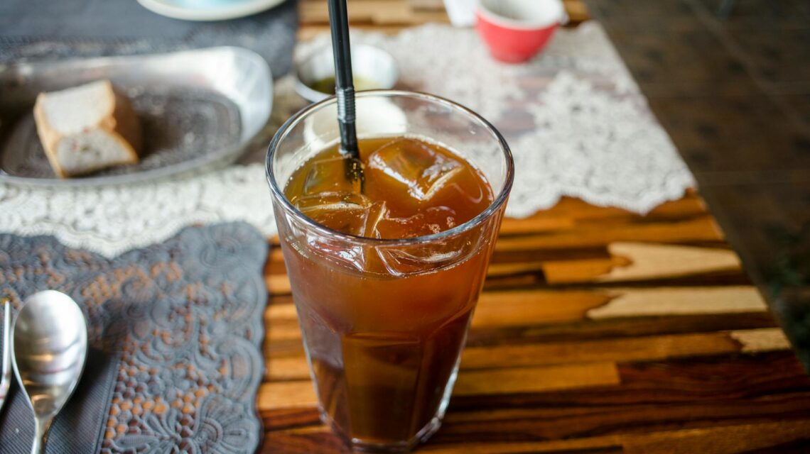 A person's bare feet propped up on a wooden railing with a condensation-beaded glass of iced tea in the foreground, overlooking a sunny garden.