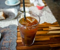 A person's bare feet propped up on a wooden railing with a condensation-beaded glass of iced tea in the foreground, overlooking a sunny garden.