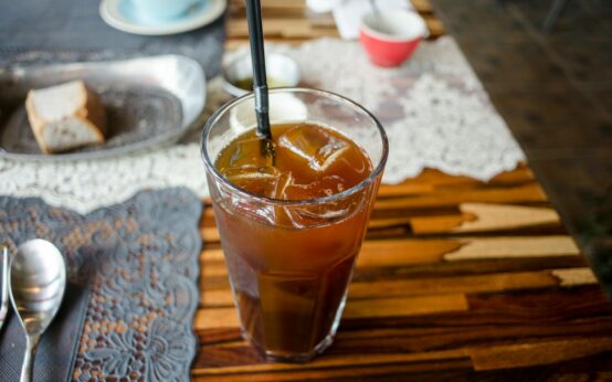 A person's bare feet propped up on a wooden railing with a condensation-beaded glass of iced tea in the foreground, overlooking a sunny garden.
