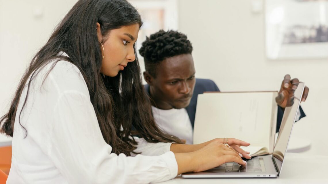 A diverse group of college students sit around a table, actively discussing a group project with laptops open.