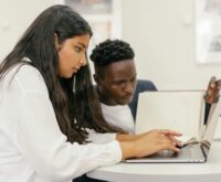 A diverse group of college students sit around a table, actively discussing a group project with laptops open.