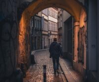 A lone traveler in a trench coat strolls down a narrow, historic cobblestone alleyway in a European city.