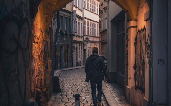 A lone traveler in a trench coat strolls down a narrow, historic cobblestone alleyway in a European city.