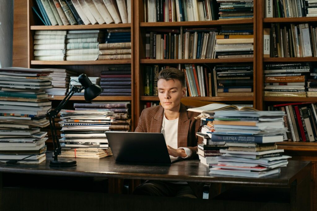 A young female student with headphones on, writing intently in a notebook in a quiet, sunlit library.