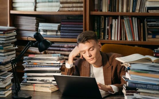 A young student with glasses focused on a book in a quiet, well-lit library.